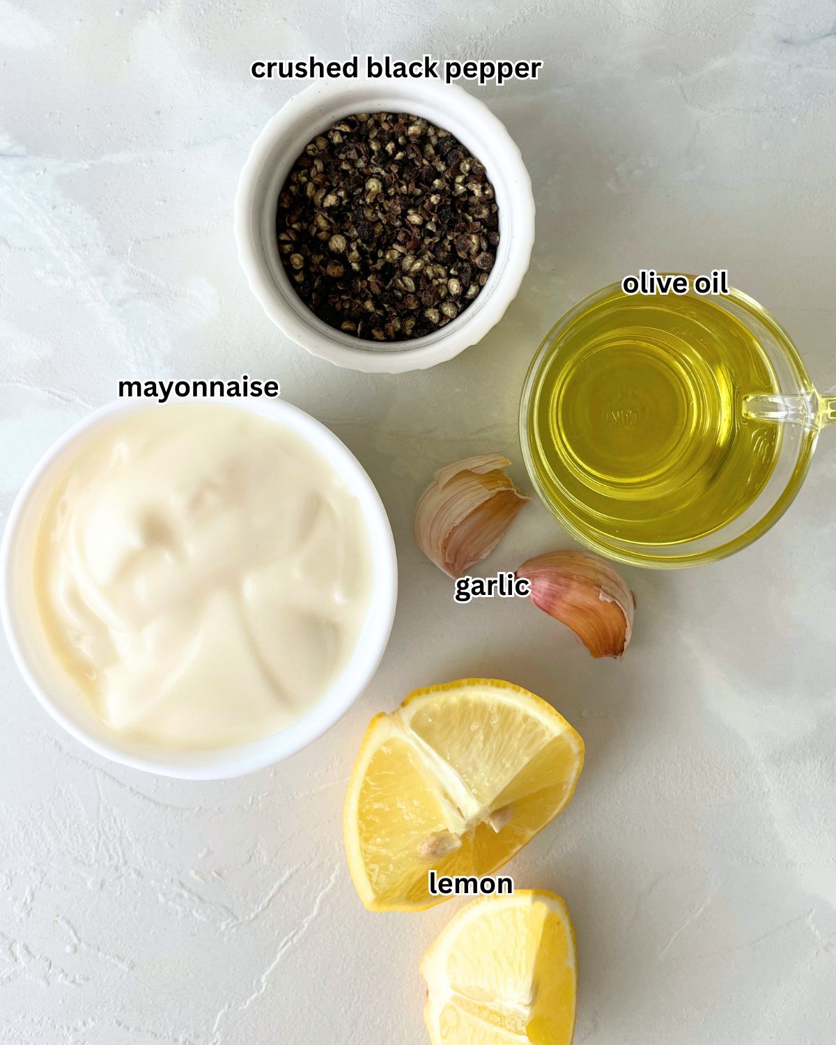 Labeled ingredients for black pepper aioli including a bowl of mayonnaise, olive oil, fresh garlic cloves, crushed black pepper in a small bowl, and lemon juice on a white surface.