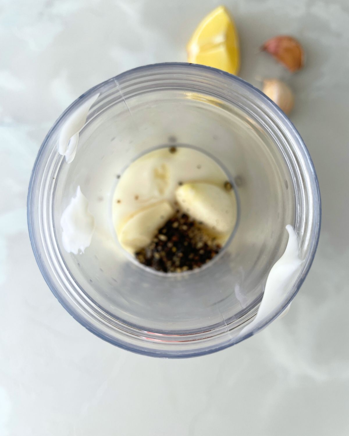 Mayonnaise, garlic cloves, crushed black pepper, lemon juice, and olive oil added to a food processor bowl, ready to be blended into black pepper aioli.