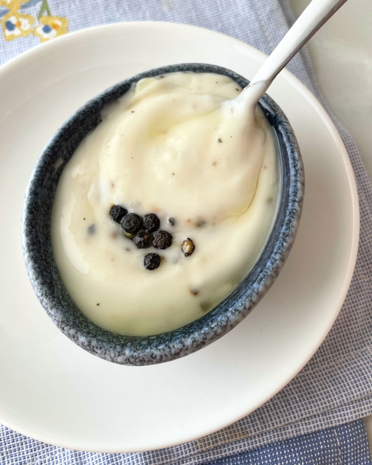 An angled view of black pepper aioli in a white bowl showing the thick, creamy texture and black pepper speckles, placed on a wooden surface.