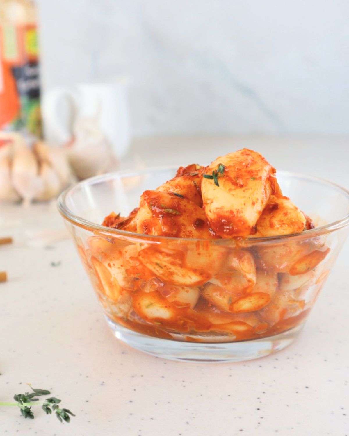 A bowl of TikTok spicy pickled garlic showing whole garlic cloves coated in glossy red Sriracha and chili flakes, with dried thyme visible throughout.