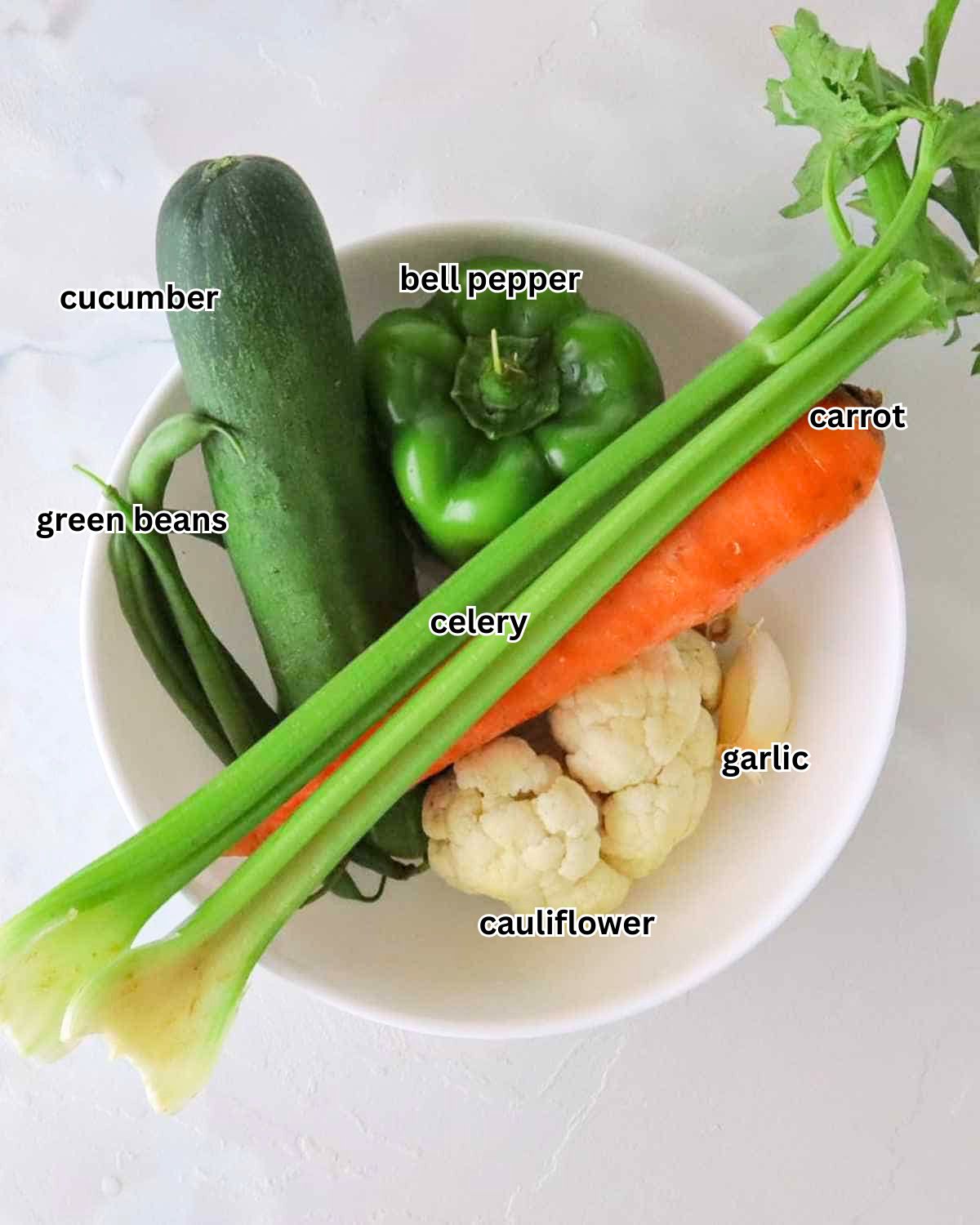 A flat lay of fresh vegetables for Persian Torshi including cauliflower florets, Persian cucumbers, a carrot, celery stalks, a green bell pepper, green beans, and whole garlic cloves on a white surface.