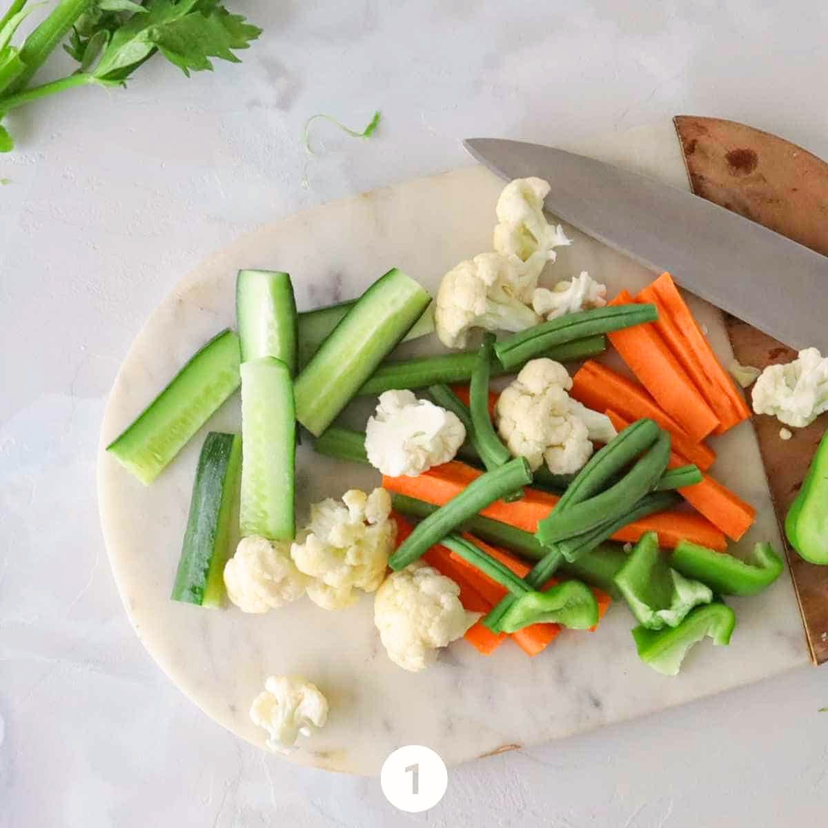 Fresh cauliflower being broken into florets and Persian cucumbers, carrots, celery, and bell pepper being chopped into bite-sized pieces on a cutting board for Persian Torshi Makhloot.